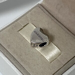 Może przedstawiać: Srebrny charm w kształcie serca z wygrawerowanym napisem "HAMBURG". Charm ma czarny detal z emalii.