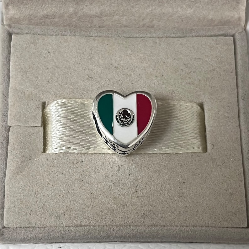 Puede incluir: Un charm de plata en forma de coraz&oacute;n con los colores de la bandera mexicana y el escudo de armas mexicano en el centro.