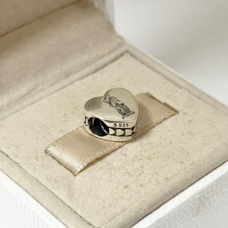 Puede incluir: Un charm plateado en forma de coraz&oacute;n con un grabado de sirena y el texto "S925". El charm tiene una banda negra alrededor del centro con un patr&oacute;n de peque&ntilde;os corazones. Descansa sobre una cinta beige dentro de una caja blanca.