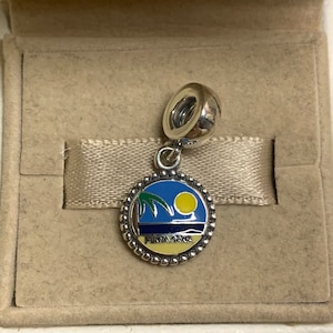 Op de afbeelding: Zilveren bedel met een blauw en geel emaille ontwerp van een palmboom en een strand met de tekst "Punta Cana" op een witte achtergrond.
