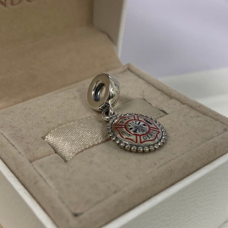 May include: Silver charm with a red and white enamel fire department logo. The logo features a fire hydrant, an axe, and a ladder. The charm is in a white box.