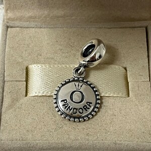 May include: A silver charm with a round, textured disc featuring the Pandora logo with a crown above the letter "O".