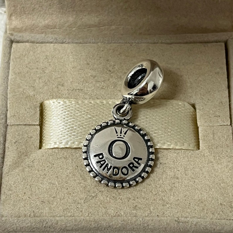 May include: A silver charm with a round, textured disc featuring the Pandora logo with a crown above the letter "O".