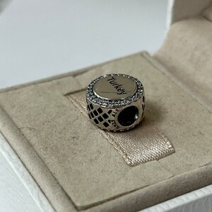 May include: A silver charm with a black and white chequered pattern and a round silver charm with the word "Turkey" in black lettering and a circle of clear stones around the edge.