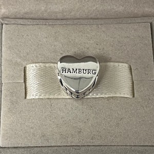 Może przedstawiać: Srebrny charm w kształcie serca z wygrawerowanym napisem "HAMBURG" z przodu. Charm znajduje się w białym pudełku.