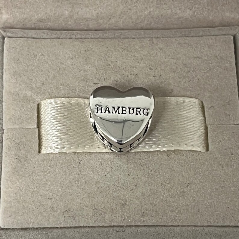 Może przedstawiać: Srebrny charm w kształcie serca z wygrawerowanym napisem "HAMBURG" z przodu. Charm znajduje się w białym pudełku.