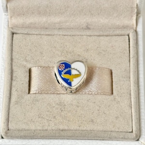 May include: A silver heart-shaped charm with blue, white, and yellow enamel. The charm features a crest with a red shield and a gold bird with outstretched wings.