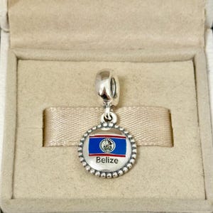 Pode incluir: Um berloque pendente de prata com um pingente circular de borda perolada. O centro exibe a bandeira nacional de Belize em esmalte azul, branco e vermelho, com a palavra Belize impressa em preto abaixo do emblema da bandeira na superfície.