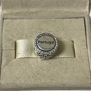 Puede incluir: Un charm de plata con un borde de cristal blanco y la palabra "Portugal" en letras negras.