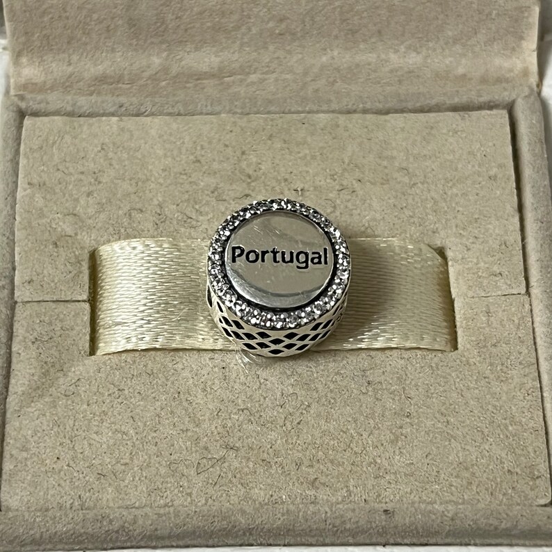 Puede incluir: Un charm de plata con un borde de cristal blanco y la palabra "Portugal" en letras negras.