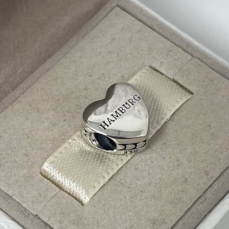 Może przedstawiać: Srebrny charm w kształcie serca z wygrawerowanym napisem "HAMBURG". Charm ma szczeg&oacute;łowy wz&oacute;r wok&oacute;ł krawędzi.