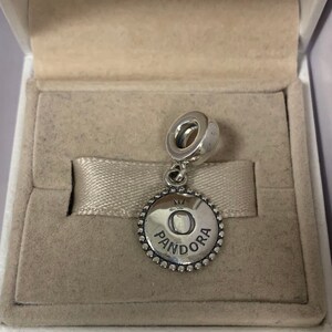 May include: A silver charm with a round pendant featuring the word "Pandora" and a crown above the letter "O".
