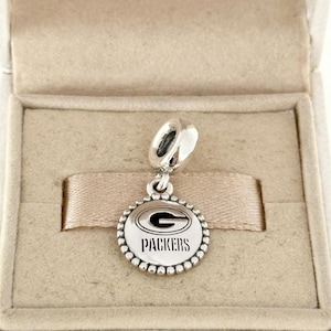 May include: A silver charm with a round pendant featuring the Green Bay Packers logo in black and white. The charm has a beaded edge and a loop for attaching to a bracelet or necklace. The charm is displayed in a beige jewelry box.