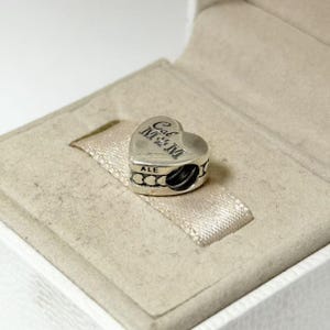 Puede incluir: Un charm plateado en forma de coraz&oacute;n con el texto grabado "Cal M&M" en la parte delantera. El charm tiene una peque&ntilde;a banda decorativa alrededor de la parte inferior con el texto "ALE". El charm se muestra dentro de una caja de joyer&iacute;a beige claro.