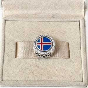May include: A silver charm with a blue and red enamel design of the Icelandic flag. The charm is surrounded by a circle of clear cubic zirconia stones.