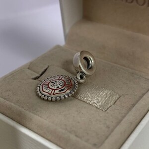 May include: Silver charm with a red and white fire department logo. The charm is in a white box with a beige interior.