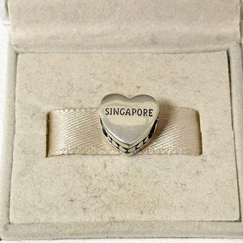 Puede incluir: Un charm plateado en forma de coraz&oacute;n con la palabra "SINGAPORE" en letras negras. El charm est&aacute; en una caja beige con una cinta de raso.