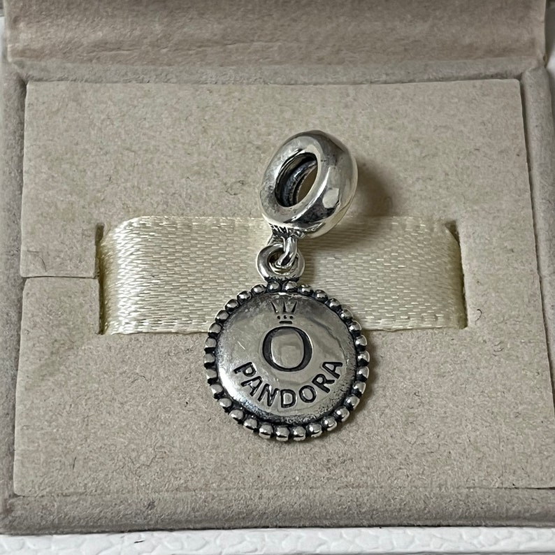 Puede incluir: Encanto de plata con un disco redondo texturizado que presenta la palabra "PANDORA" y una corona sobre la letra "O".