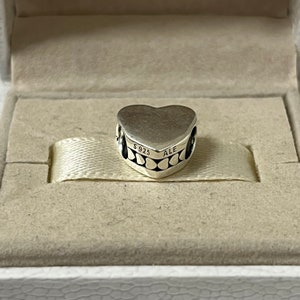 May include: A silver heart-shaped charm with a detailed design around the edge. The charm is engraved with "S925 ALE" on the back.