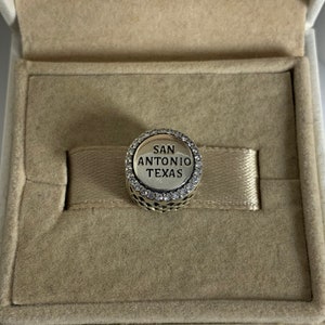 Puede incluir: Un charm plateado con un borde de cristal transparente y el texto "SAN ANTONIO TEXAS" en letras negras.