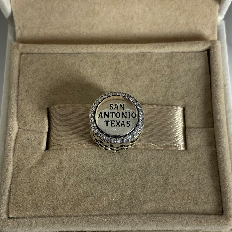 Puede incluir: Un charm plateado con un borde de cristal transparente y el texto "SAN ANTONIO TEXAS" en letras negras.