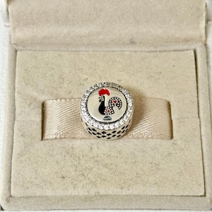 Könnte beinhalten: Ein runder Silberanhänger mit einem schwarzen und roten Hahn-Design. Der Anhänger ist mit klaren Kristallen am Rand besetzt und wird in einer beigen Box präsentiert.