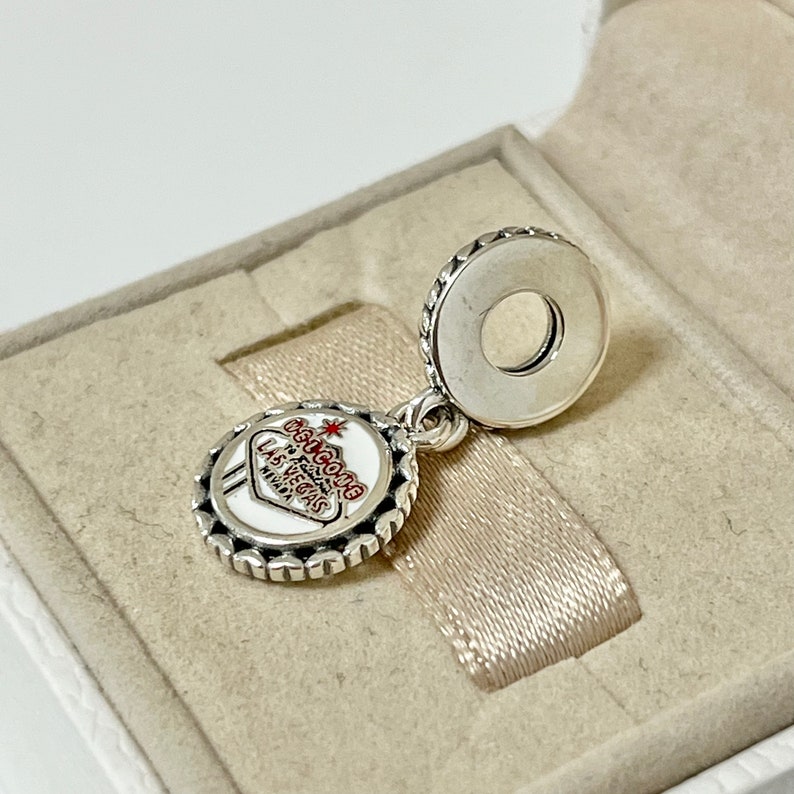 Puede incluir: Encanto de plata con un letrero de esmalte blanco que dice "Bienvenido a Las Vegas" con una estrella roja y letras rojas. El encanto est&aacute; en una caja blanca con una cinta plateada.