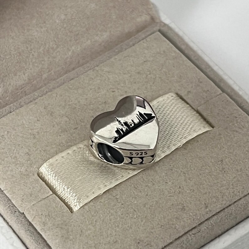 Może przedstawiać: Srebrny charm w kształcie serca z sylwetką miasta w środku. Charm ma mały otw&oacute;r do przymocowania do bransoletki. Tył charmu jest wytłoczony "S 925".