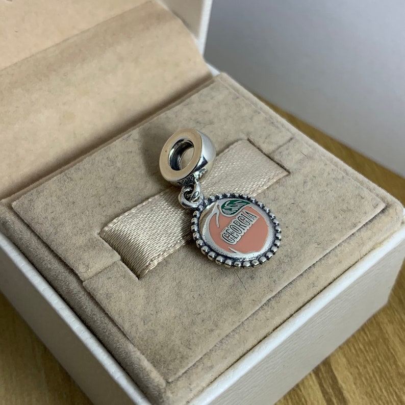May include: A silver charm in the shape of a peach with the word "Georgia" written on it. The peach is pink and has a green leaf.