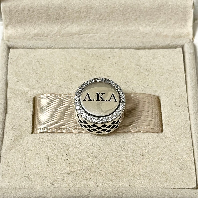 May include: A silver charm with a black and white checkered pattern and a circle with clear stones around the edge. The circle has the letters "A.K.A." in black lettering.