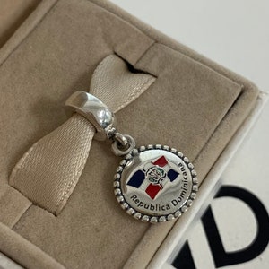May include: Silver charm with a round, enamel-painted design featuring the flag of the Dominican Republic. The charm has the text "Republica Dominicana" around the edge.