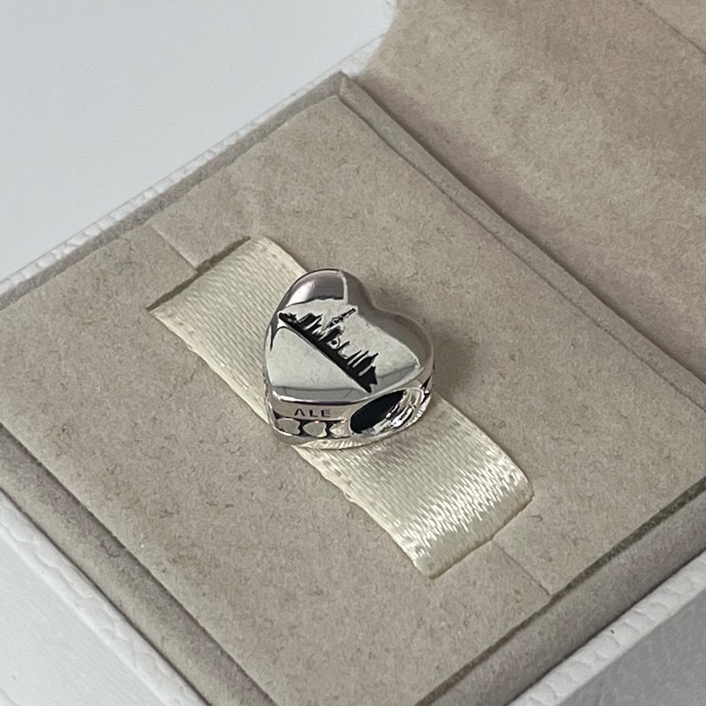 Może przedstawiać: Srebrny charm w kształcie serca z sylwetką miasta i wygrawerowanym napisem "ALE" na boku.