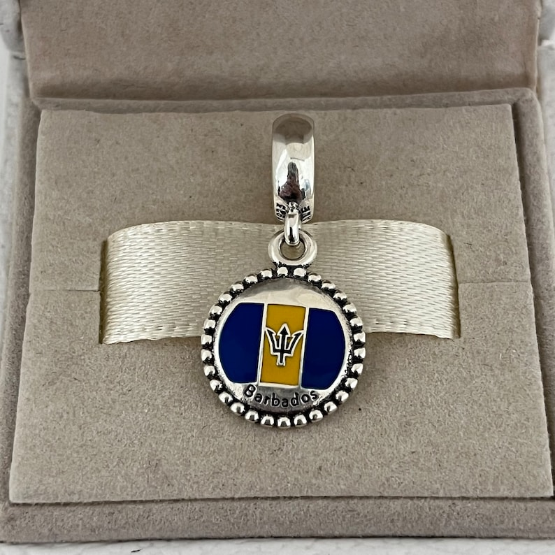 Op de afbeelding: Zilveren bedel met een blauw en geel emaille ontwerp van de vlag van Barbados. De bedel heeft een parelrand en het woord "Barbados" staat onder de vlag.