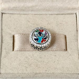 May include: A silver charm with a blue and white enamel design of Stitch from Lilo & Stitch. The charm is surrounded by a circle of clear cubic zirconia stones.