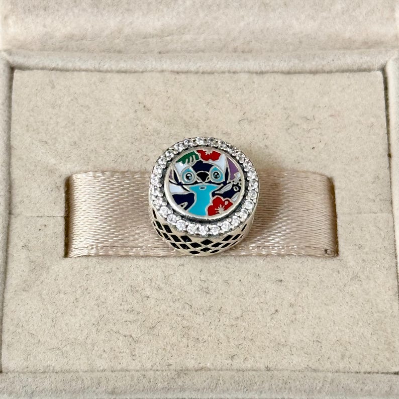 May include: A silver charm with a blue and white enamel design of Stitch from Lilo & Stitch. The charm is surrounded by a circle of clear cubic zirconia stones.