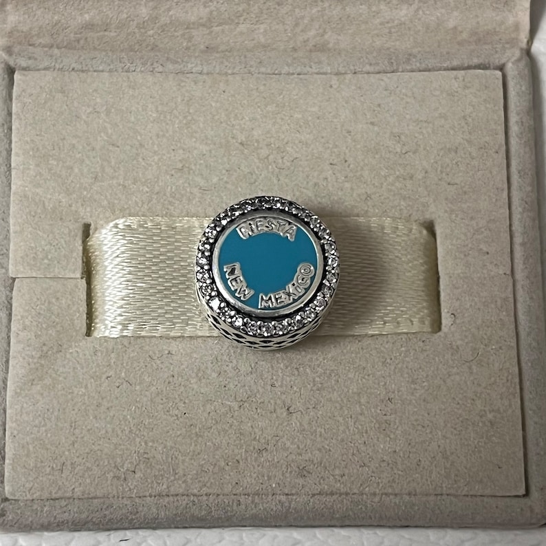 May include: A silver charm with a blue enamel circle that says "Fiesta New Mexico" in white lettering. The charm is surrounded by a ring of clear cubic zirconia stones.