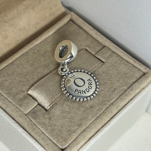 May include: Silver charm with a round pendant featuring the word "Pandora" and a crown above the letter "O".