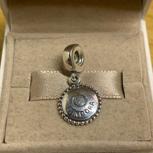 May include: A silver charm with a round, beaded edge. The charm features a circular design with the word "PANDORA" and a crown.