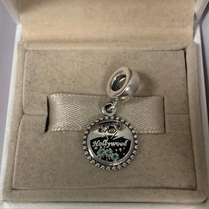 May include: Silver charm featuring a black and white enamel design of the Hollywood sign with palm trees and fireworks. The charm is presented in a white box with a light brown lining.