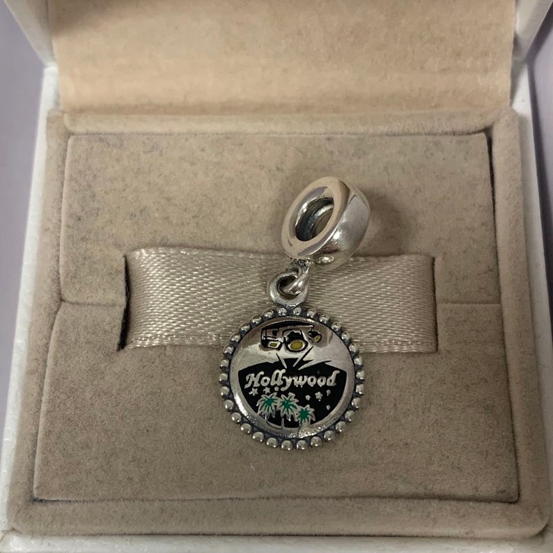 May include: Silver charm featuring a black and white enamel design of the Hollywood sign with palm trees and fireworks. The charm is presented in a white box with a light brown lining.