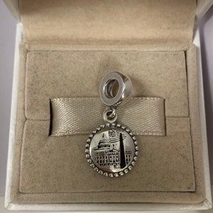 Puede incluir: Un charm de plata con una imagen detallada del Capitool de los Estados Unidos con una bandera estadounidense en primer plano. El charm es redondo y tiene un borde de cuentas.