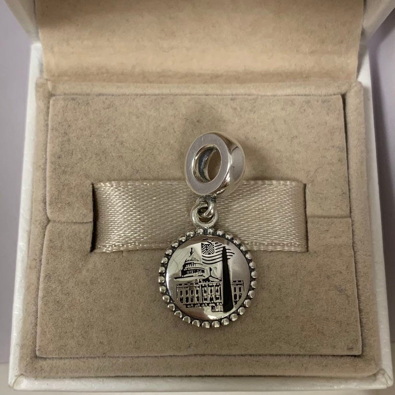 Puede incluir: Un charm de plata con una imagen detallada del Capitool de los Estados Unidos con una bandera estadounidense en primer plano. El charm es redondo y tiene un borde de cuentas.
