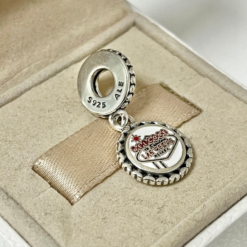 Puede incluir: Encanto de plata con un letrero de Las Vegas de esmalte rojo y blanco que dice "Welcome to Fabulous Las Vegas Nevada". La parte posterior del encanto tiene la inscripci&oacute;n "S925 ALE".