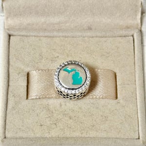 Puede incluir: Un charm de plata con un contorno turquesa del estado de Michigan, rodeado de cristales transparentes. El charm se presenta en una caja de joyería beige con una cinta.