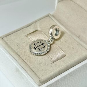 May include: A silver charm with a round pendant featuring a black scale design, surrounded by a beaded border. The charm is attached to a silver loop, and is displayed inside a white jewelry box.