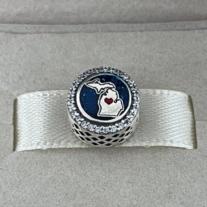 Puede incluir: Un charm de plata con un contorno de esmalte azul y blanco del estado de Michigan con un corazón rojo en el centro. El charm está rodeado por un círculo de piedras de circonita cúbica transparentes.