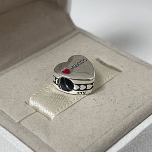 Puede incluir: Encanto de plata en forma de coraz&oacute;n con las palabras "Me encanta M&eacute;xico" y un coraz&oacute;n rojo en &eacute;l. El encanto tiene un borde negro con peque&ntilde;os corazones alrededor. El encanto est&aacute; en una caja blanca.
