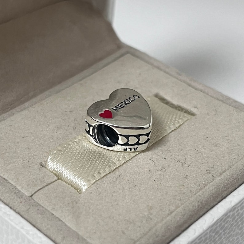Puede incluir: Encanto de plata en forma de coraz&oacute;n con las palabras "Me encanta M&eacute;xico" y un coraz&oacute;n rojo en &eacute;l. El encanto tiene un borde negro con peque&ntilde;os corazones alrededor. El encanto est&aacute; en una caja blanca.