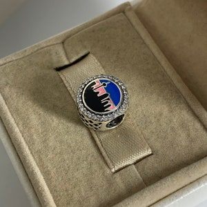 Puede incluir: Un charm de plata con un dise&ntilde;o de ciudad en blanco y negro, acentuado con esmalte rosa y azul. El charm est&aacute; rodeado por un c&iacute;rculo de piedras de circonita c&uacute;bica transparentes.