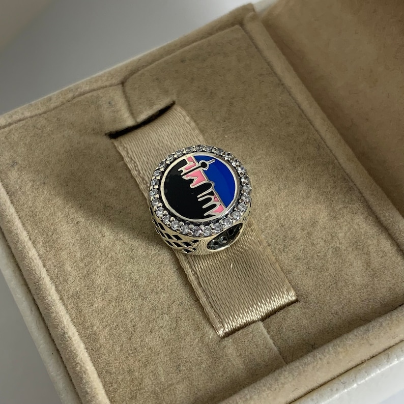 Puede incluir: Un charm de plata con un dise&ntilde;o de ciudad en blanco y negro, acentuado con esmalte rosa y azul. El charm est&aacute; rodeado por un c&iacute;rculo de piedras de circonita c&uacute;bica transparentes.